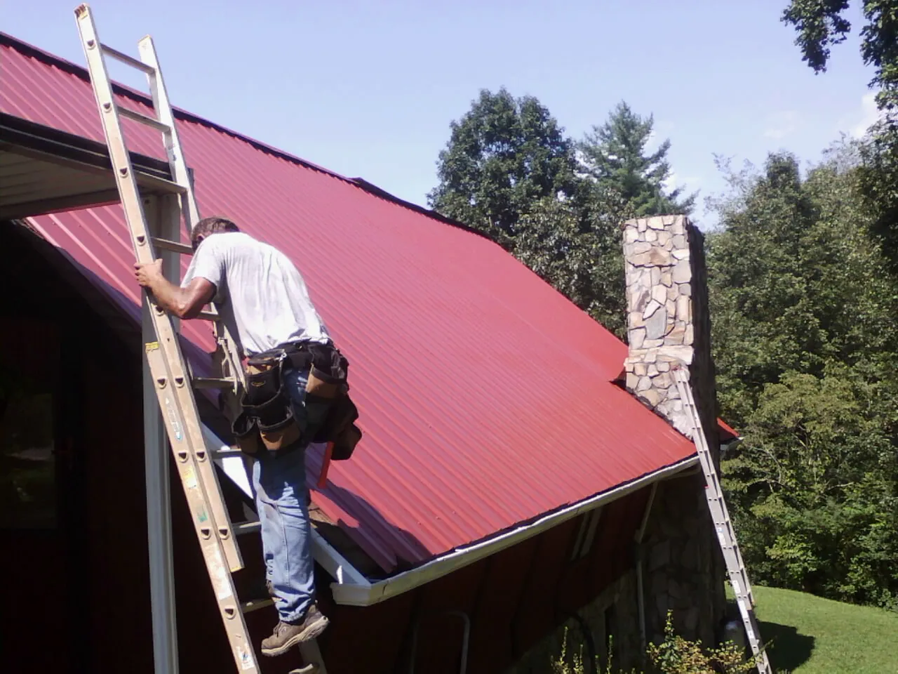 Roofer on a ladder beside a red metal roof and stone chimney — Soffit & Fascia Repair by Roofing Brothers in Stuarts Draft